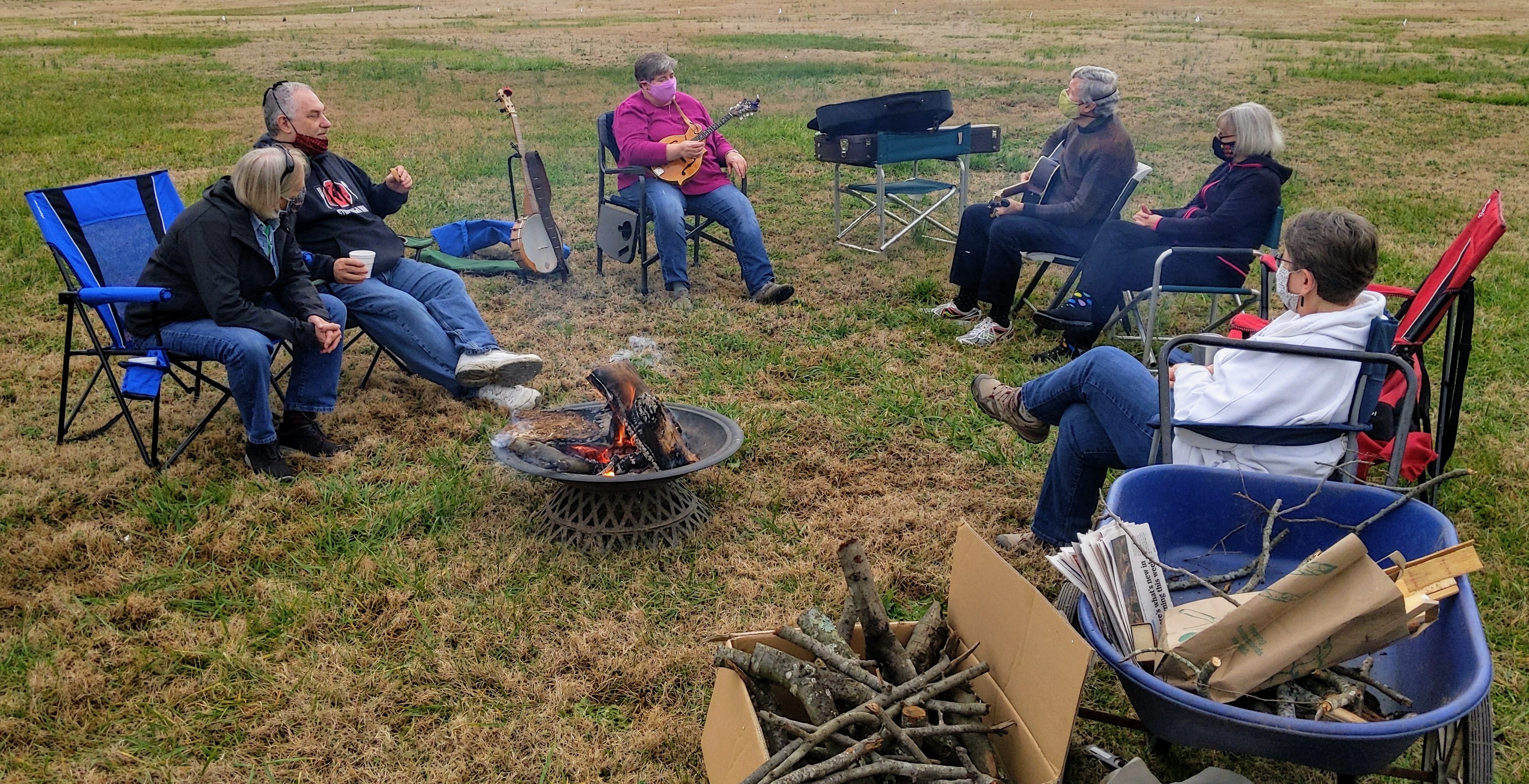 Group sitting around a fire pit in lawn chairs, some with musical instruments on their laps.