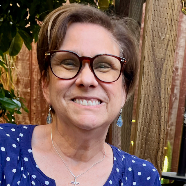 Carolyn in glasses and blue blouse, smiling with a fence and greenery behind her