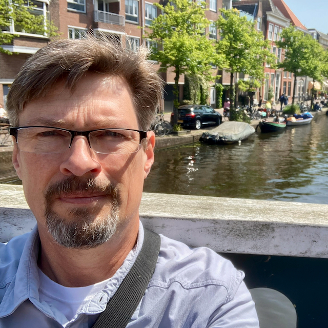 Headshot of Paul with a tight beard and mustache on a bridge with a river and some boats behind him.