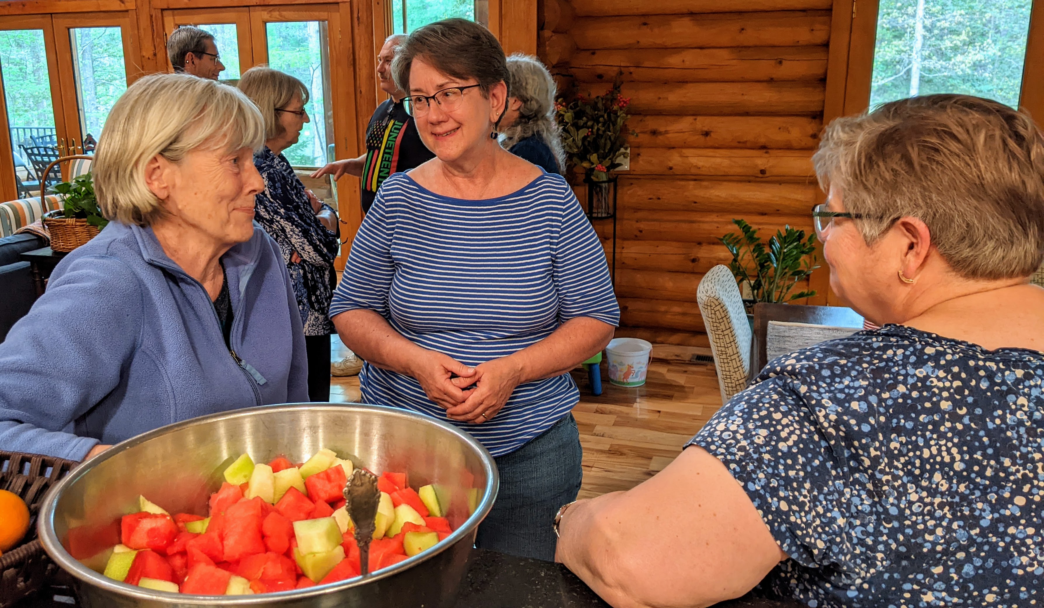 Three women inside talkin with each other, beside a large bowl of fruit salad.