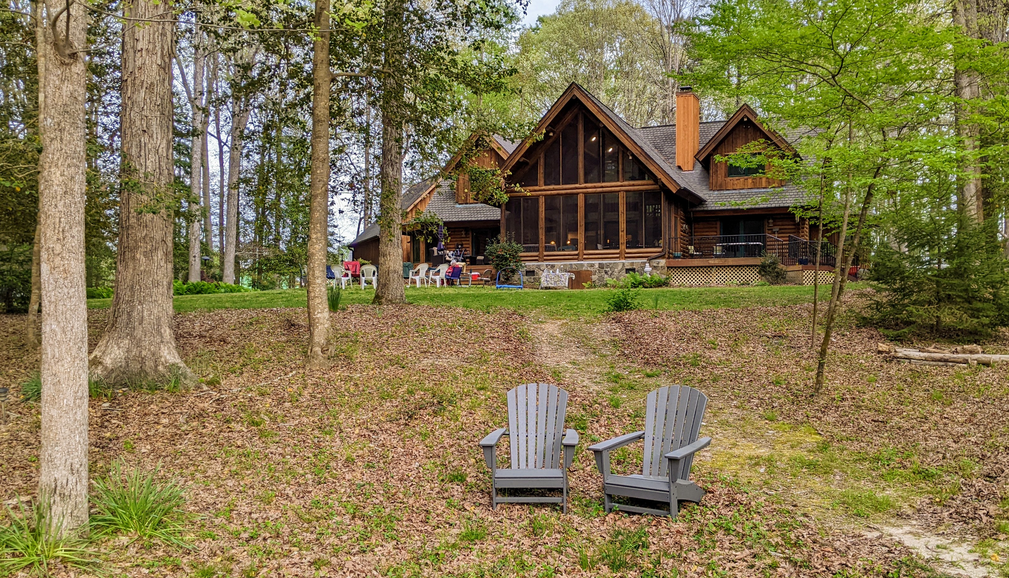 A large log cabin with cathedral windows overlooks a yard and trees with two chairs out front. 