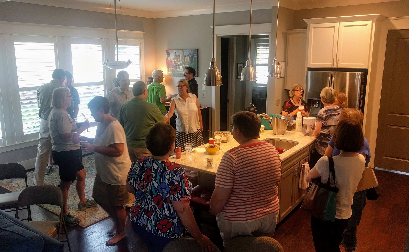 Various groups of folks standing in kitchen area holding drinks and food, talking.